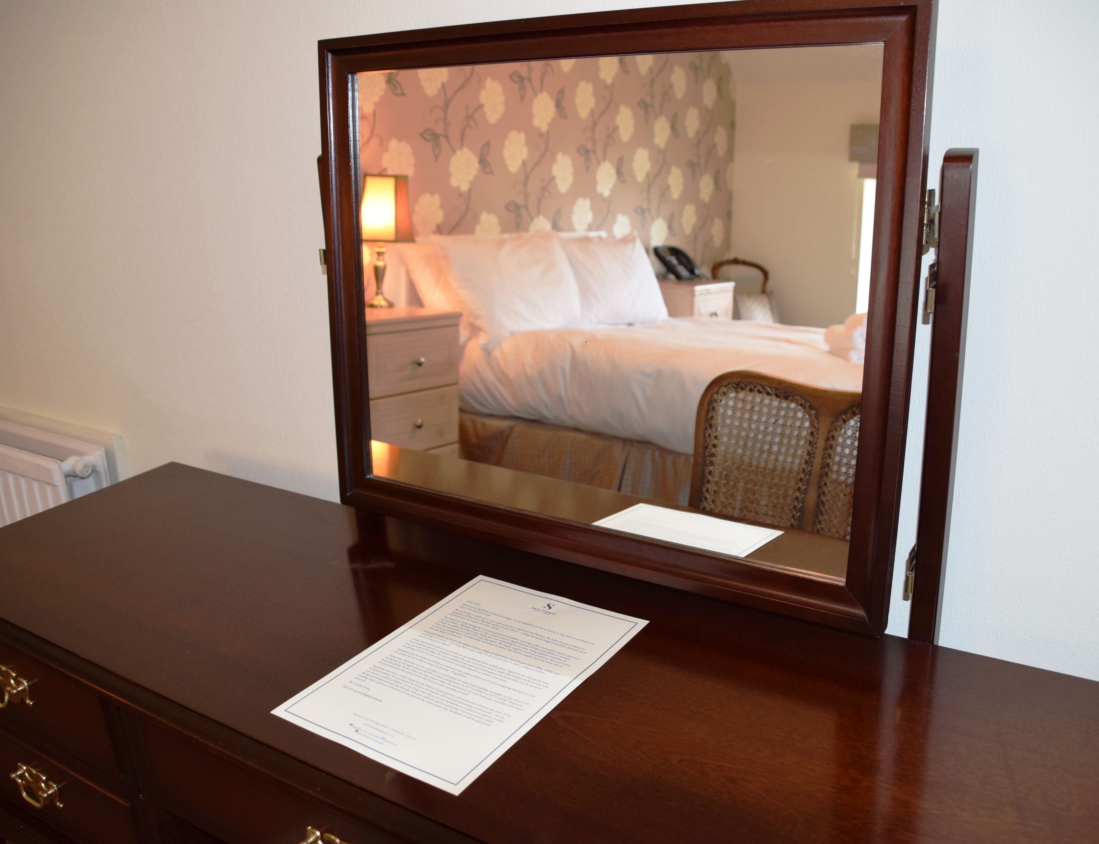 A wooden dresser with a mirror reflecting a bedroom. The room has a bed, a nightstand, and a chair. There is a lamp on the nightstand.