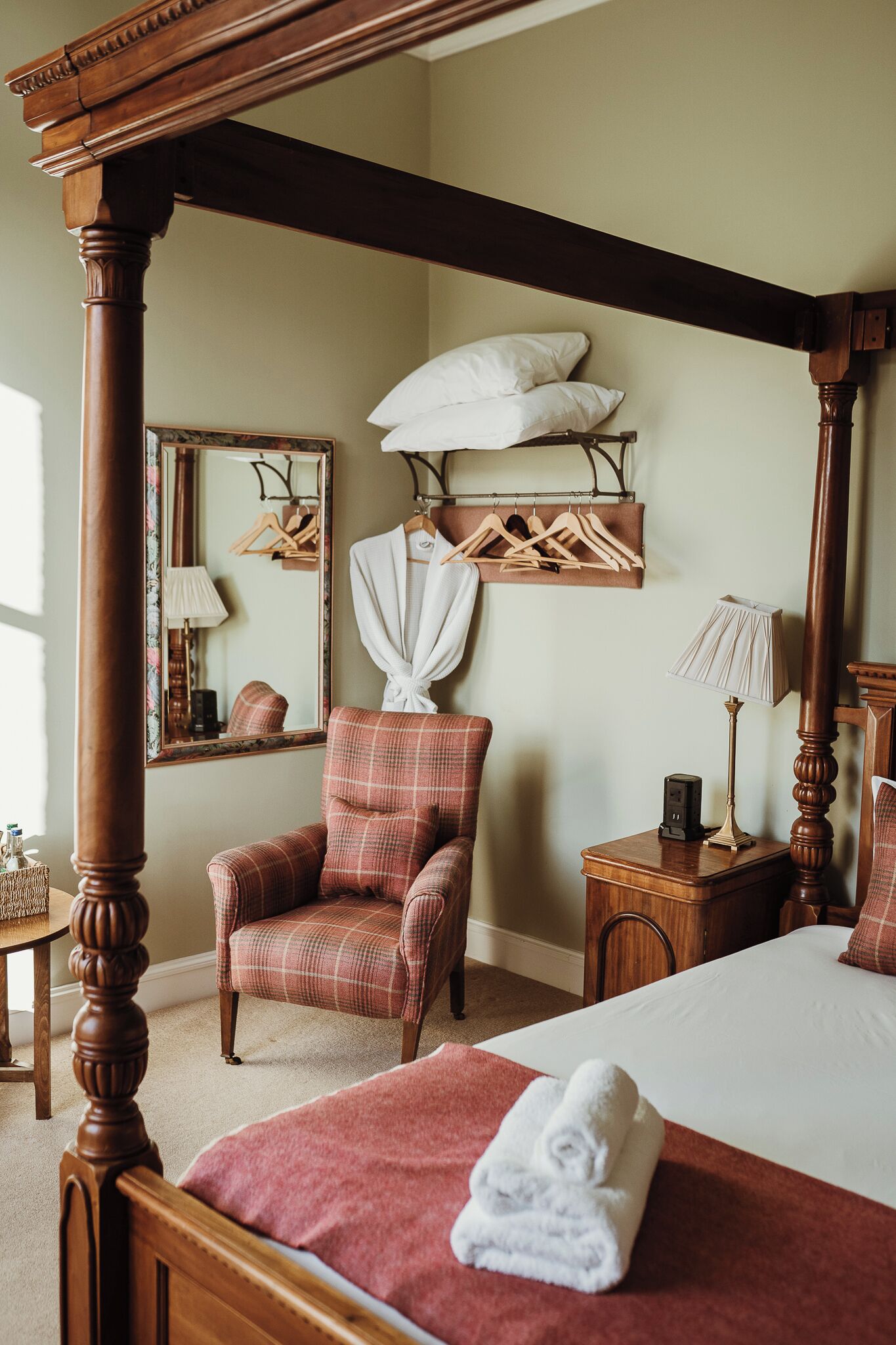 A four-poster bed with a red bedspread, a plaid armchair, a bedside table with a lamp, and a mirror on the wall.