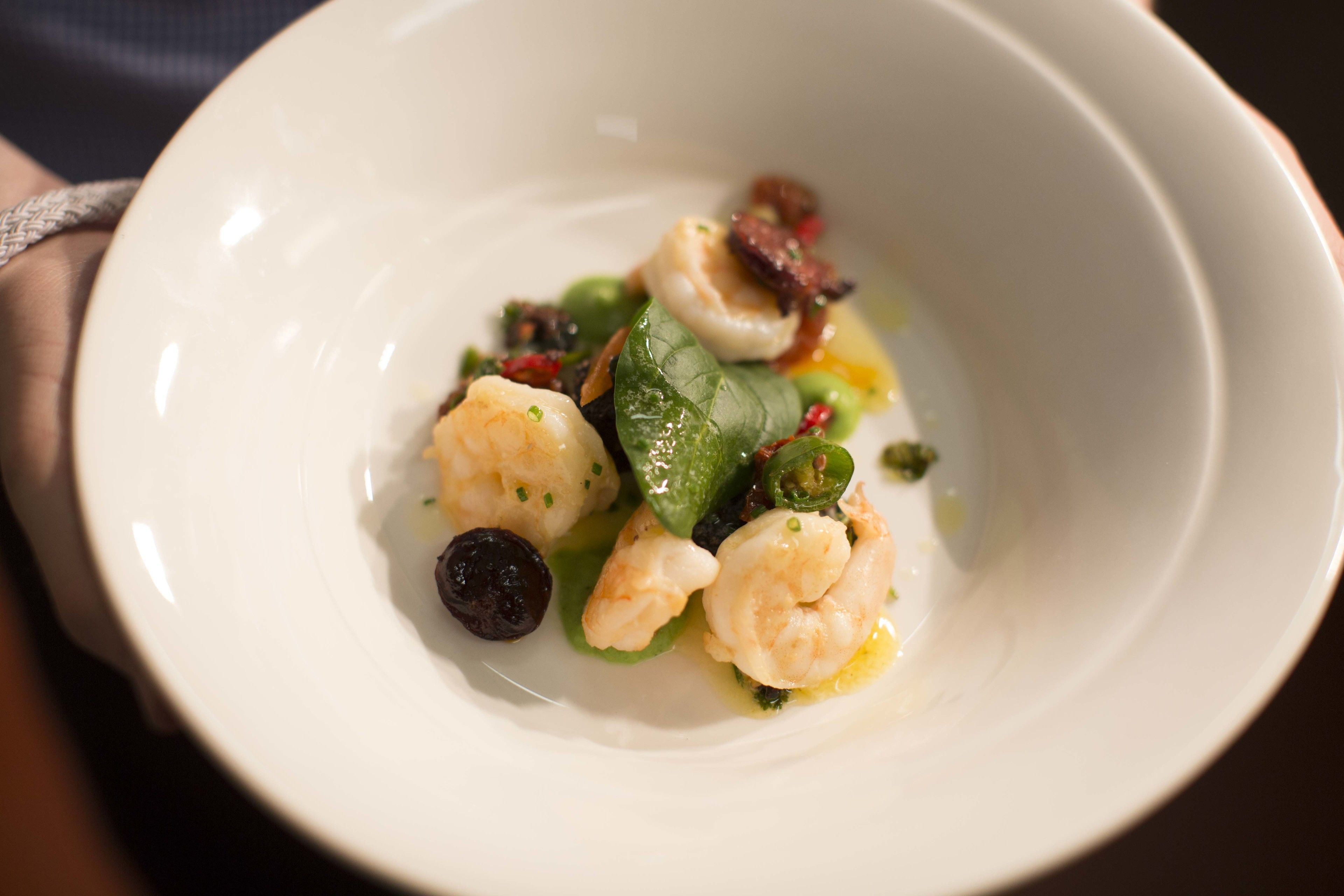 Una ciotola bianca con un piatto di pesce che include gamberi, verdure a foglia verde e un elemento sferico scuro, probabilmente una verdura.
