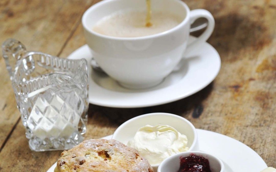 Una mesa con una taza de té, un bollo, un recipiente pequeño de mermelada y un vaso de leche.