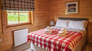 Une chambre dans une cabane en bois, avec une couverture à carreaux sur le lit, un radiateur, une fenêtre donnant sur la verdure et un tableau encadré accroché au mur.