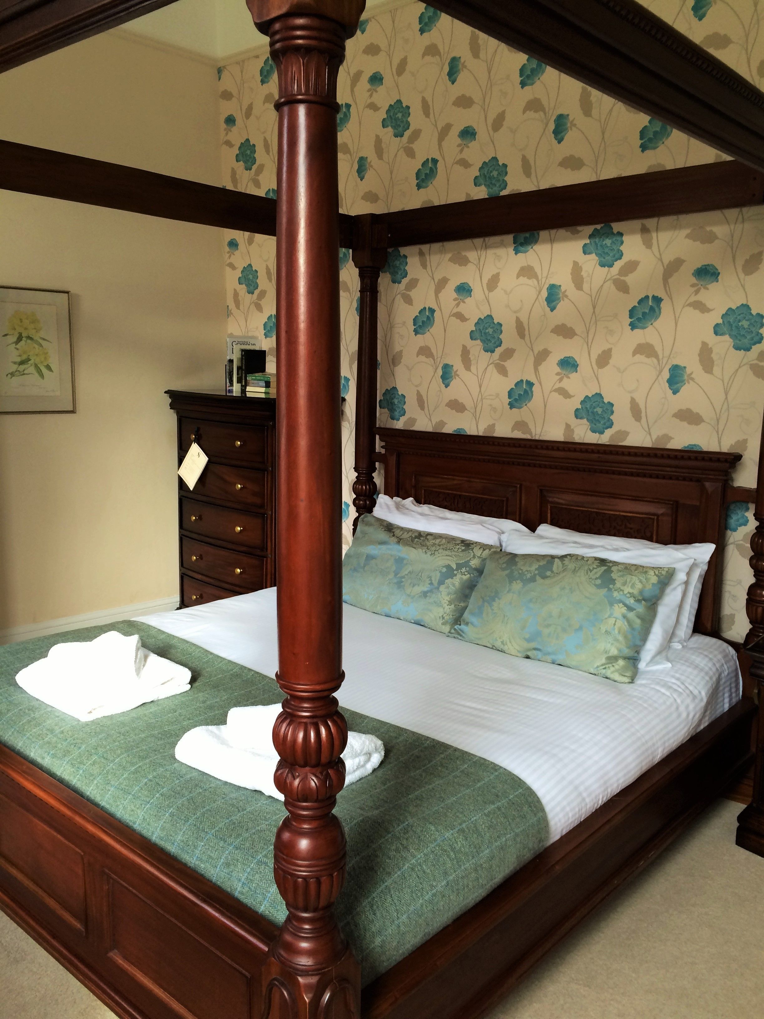 A four-poster bed with a green bedspread and white linens, a wooden nightstand, and a dresser with a floral wallpaper background.