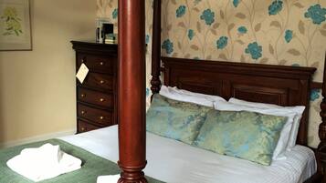 A four-poster bed with a green bedspread and white linens, a wooden nightstand, and a dresser with a floral wallpaper background.