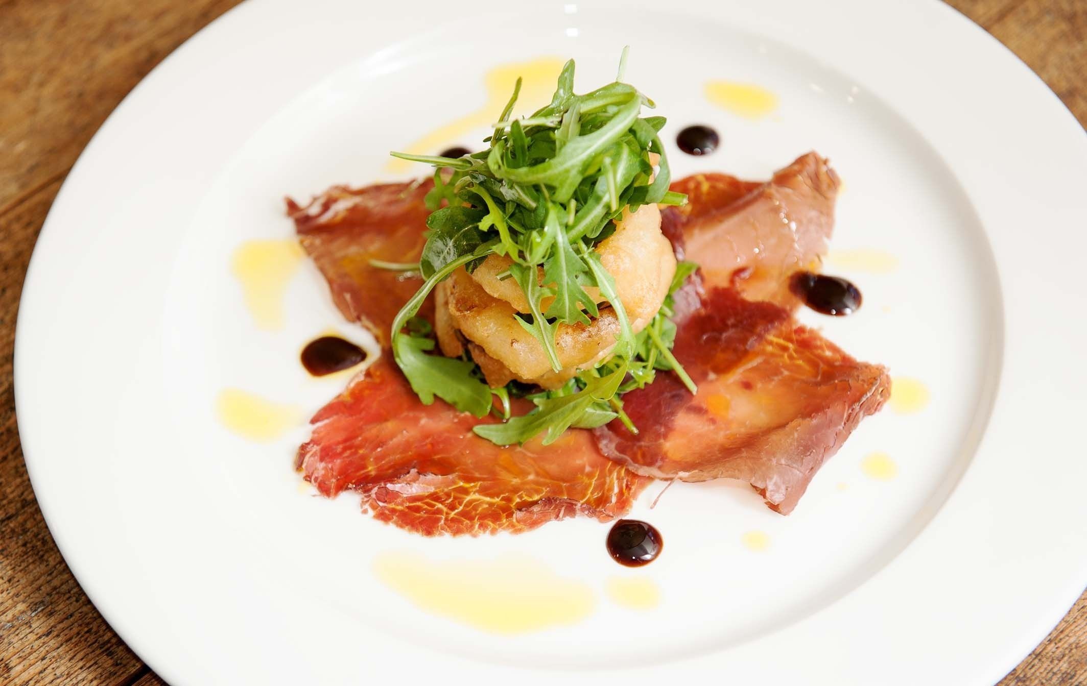 A plate with a dish of prosciutto, arugula, and a small piece of meat, possibly chicken, with a drizzle of sauce.