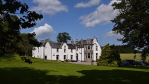 Un bâtiment traditionnel de couleur blanche, doté de nombreuses cheminées et d’un toit à forte pente, entouré d’une pelouse bien entretenue et d’arbres.