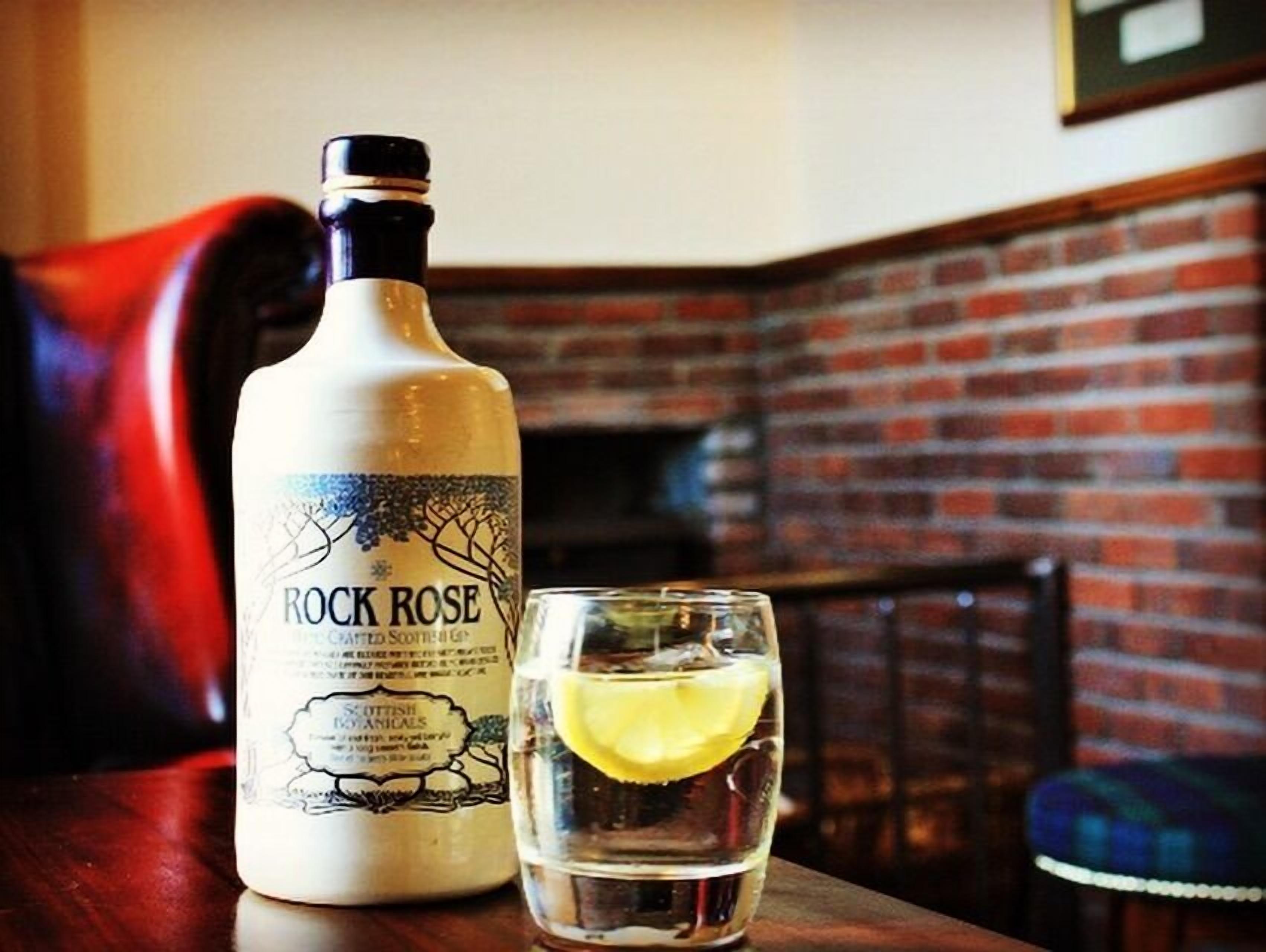 Una botella de ginebra Rock Rose y un vaso con una rodaja de limón sobre una mesa de madera.