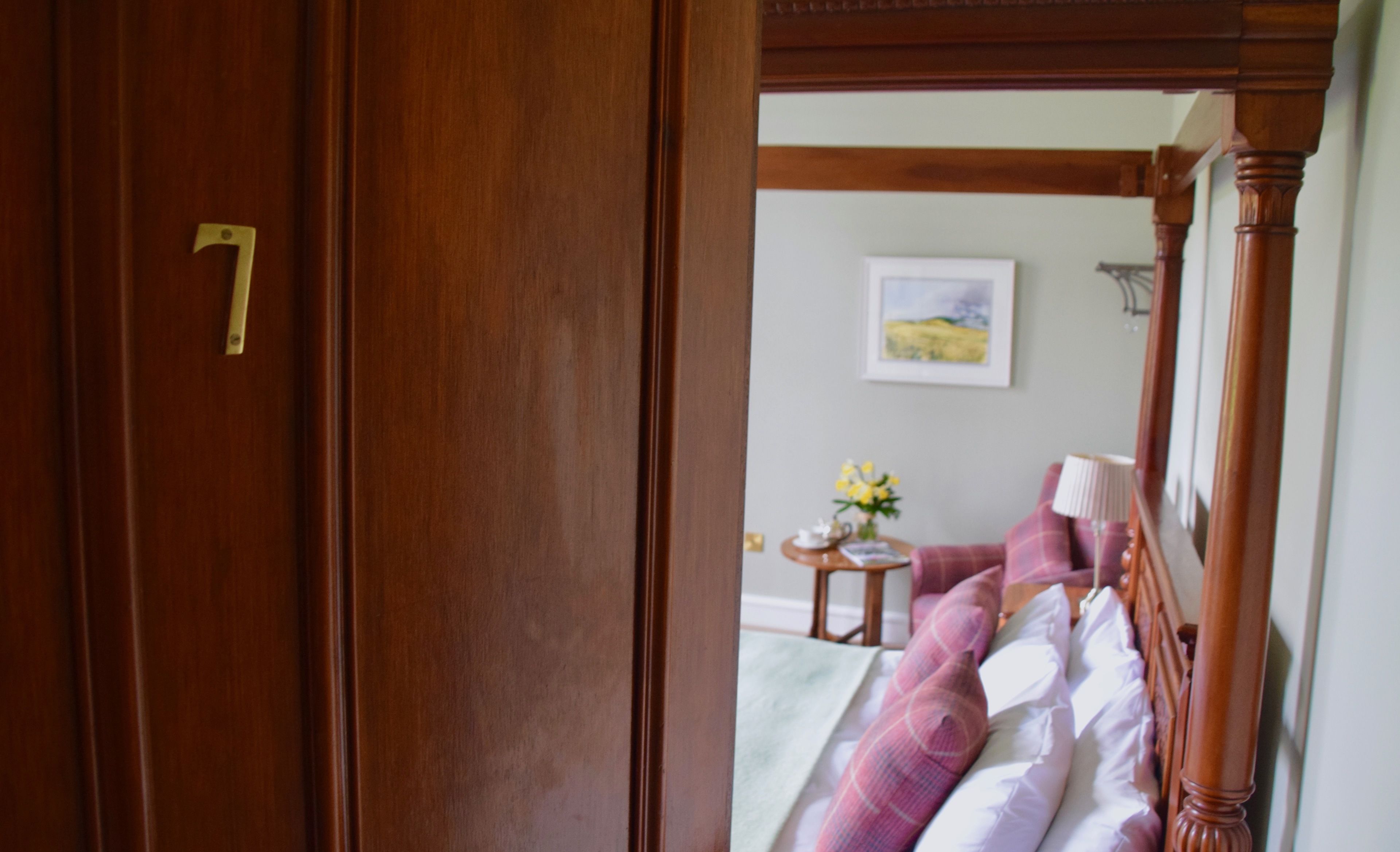 A wooden door with a brass number plate, leading to a room with a bed, a small table with a vase of flowers, and a framed picture on the wall.