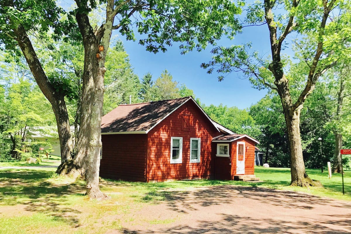 A small, single-story red wooden cabin with a sloped roof, surrounded by trees and a clear sky.