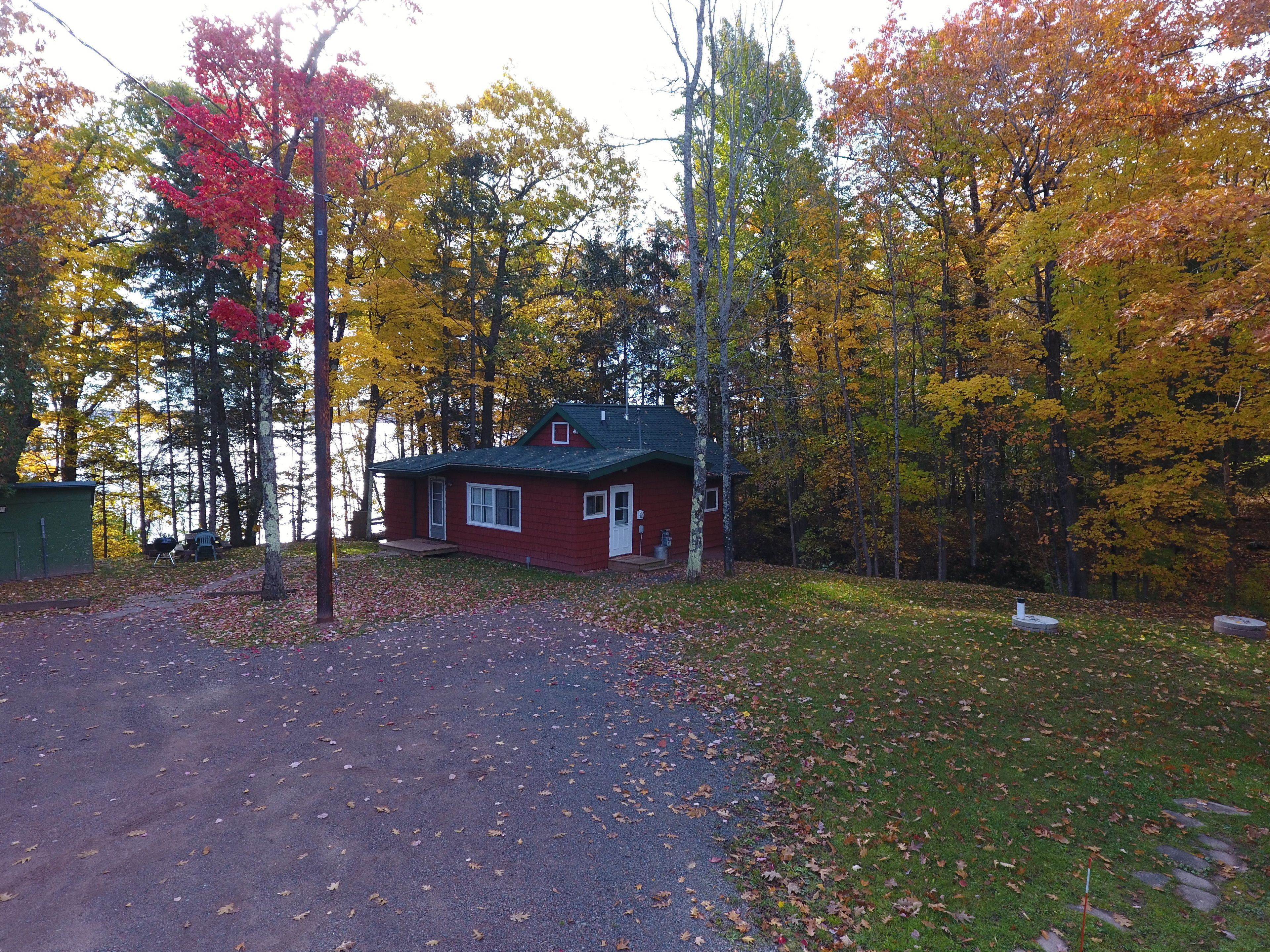 A red cabin with a green roof, surrounded by autumn foliage and a lake.