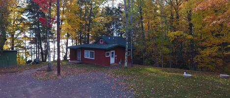 Une cabane rouge avec un toit vert, entourée d’un feuillage automnal et bordée par un lac.