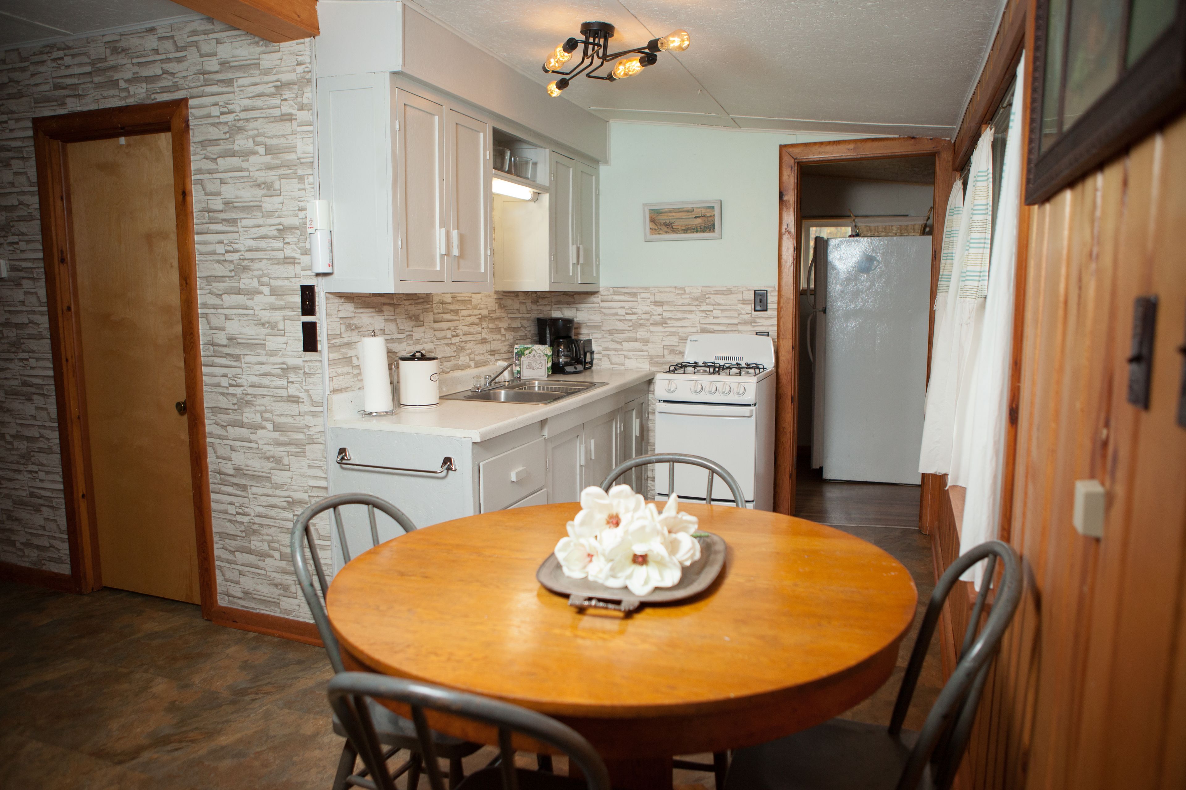 Cocina con gabinetes blancos, estufa y refrigerador. Hay una mesa de comedor con sillas y una bandeja de flores blancas.