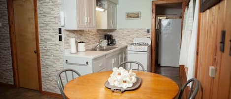 Cocina con gabinetes blancos, estufa y refrigerador. Hay una mesa de comedor con sillas y una bandeja de flores blancas.