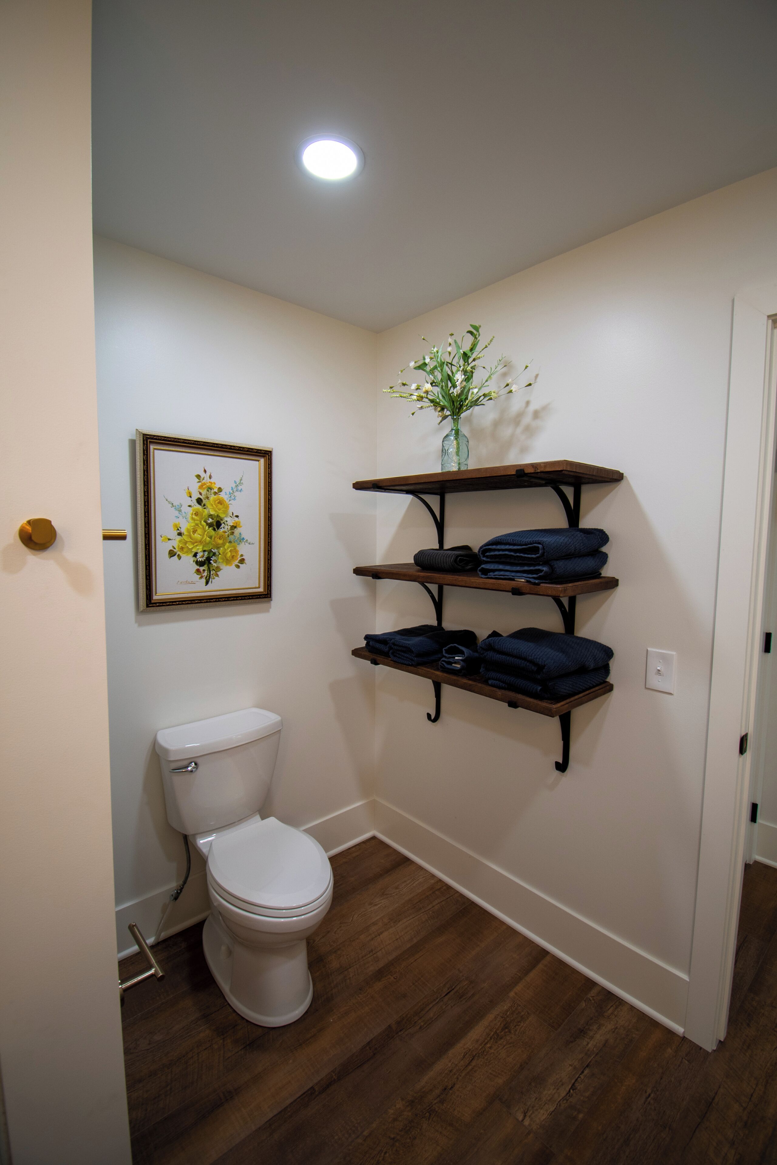 A bathroom with a toilet, wooden shelves, and a framed floral artwork.