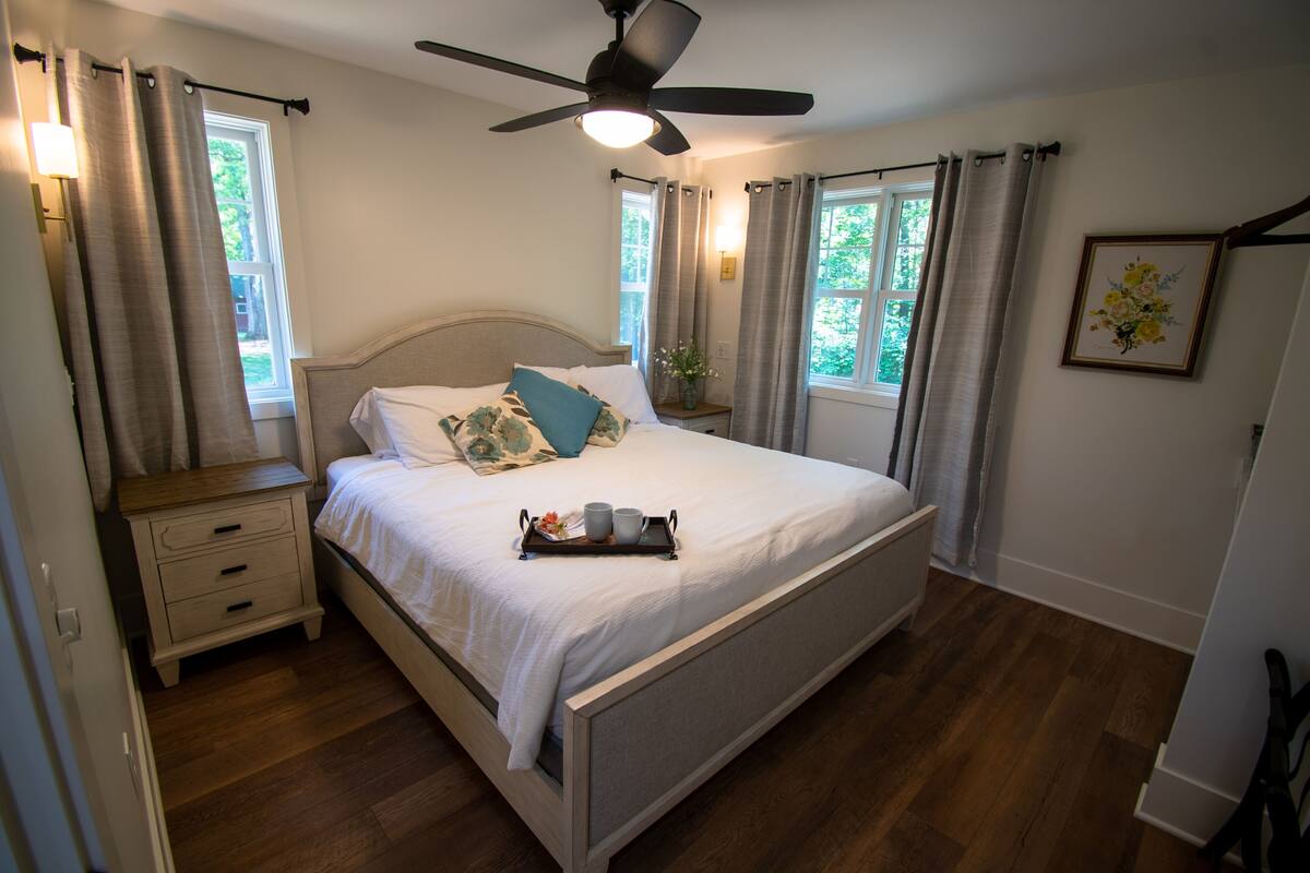 A bedroom with a bed, a nightstand, a ceiling fan, and two windows with curtains.