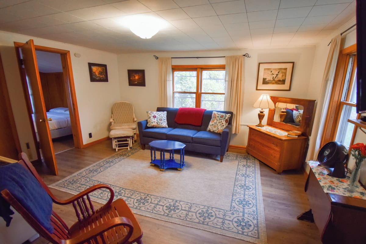A cozy living room with a blue sofa, a wooden chair, a small table, and a dresser. There is a bed in the adjacent room, and a window with curtains.
