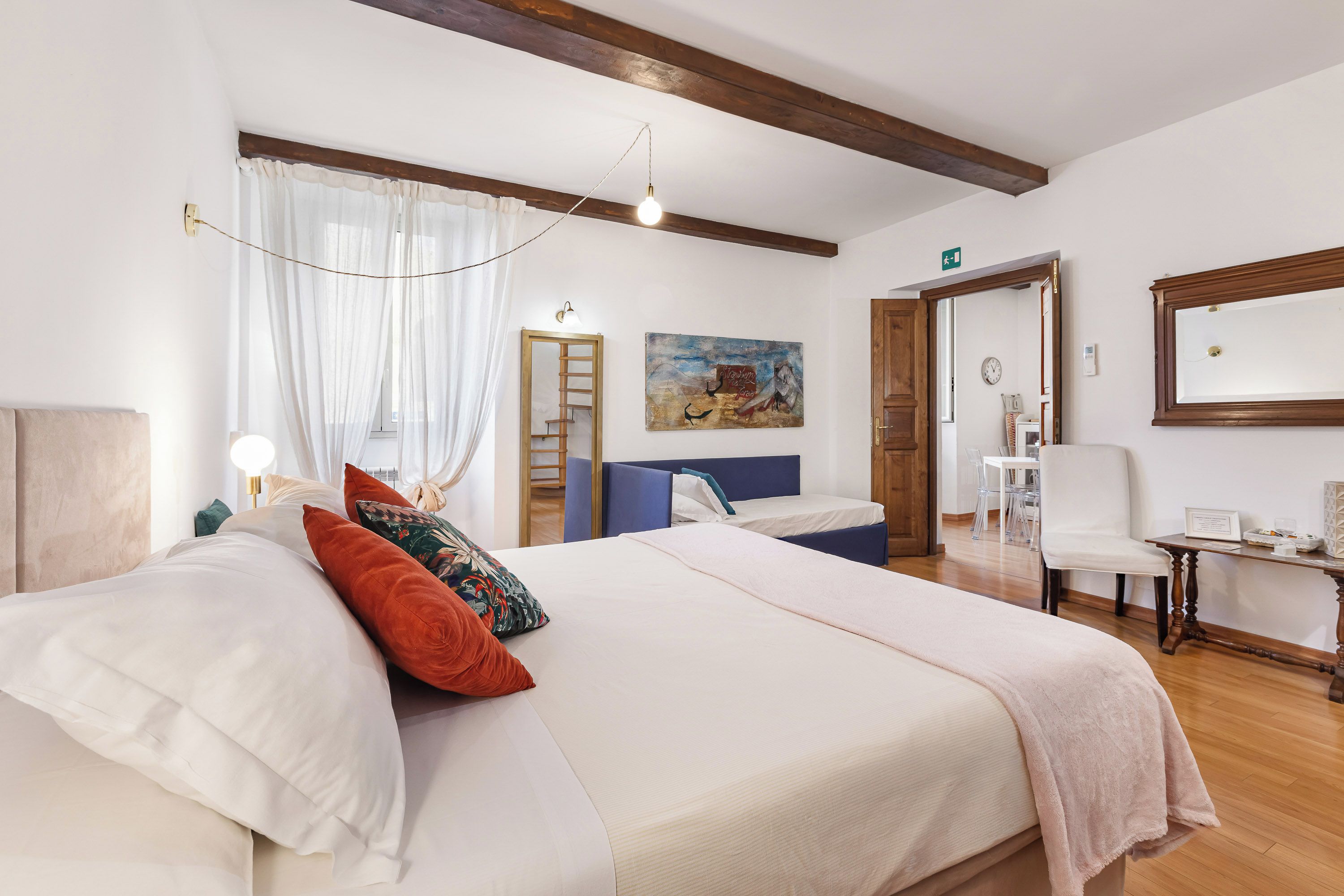 Une chambre d’hôtel avec un lit, un bureau, une chaise, un tableau et une porte donnant sur une autre pièce.