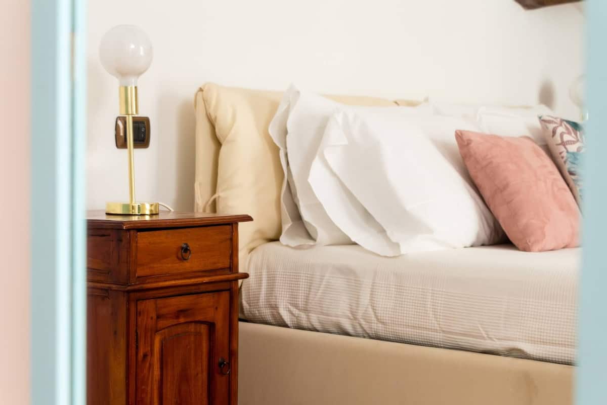 A bedroom with a wooden nightstand, a lamp, a bed with white and pink pillows, and a beige wall.