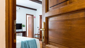 A wooden door with a brass handle, a bed with a blue blanket, a television on a stand, and a wooden cabinet in a room with white walls and wooden beams. - Torripa Group - Trastevere (Rome)