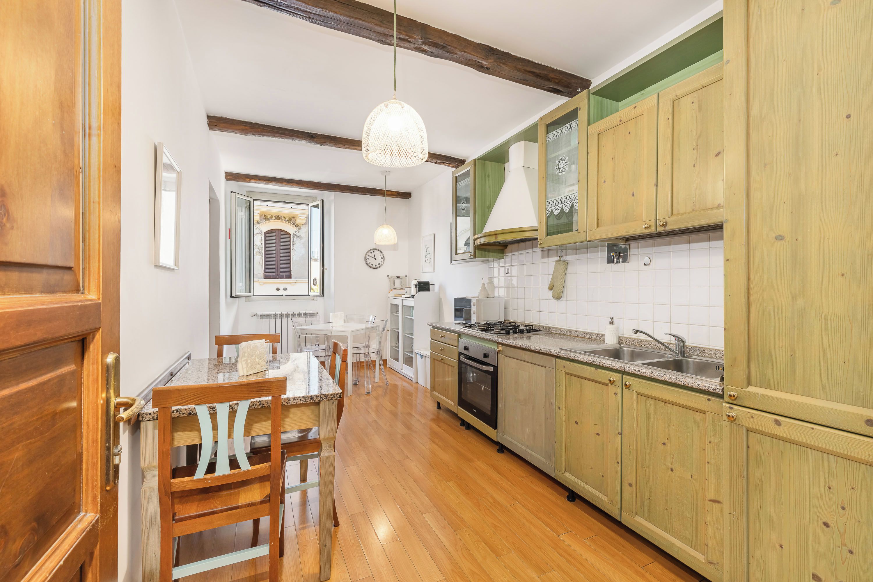 A kitchen with wooden cabinets, a dining table with chairs, a hanging light fixture, and a clock on the wall.