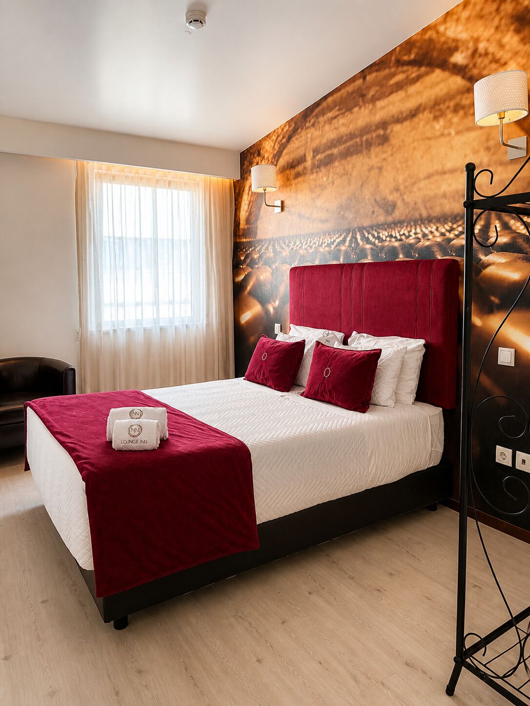 A hotel room with a large bed, a red headboard, a wall-mounted lamp, and a window with sheer curtains.