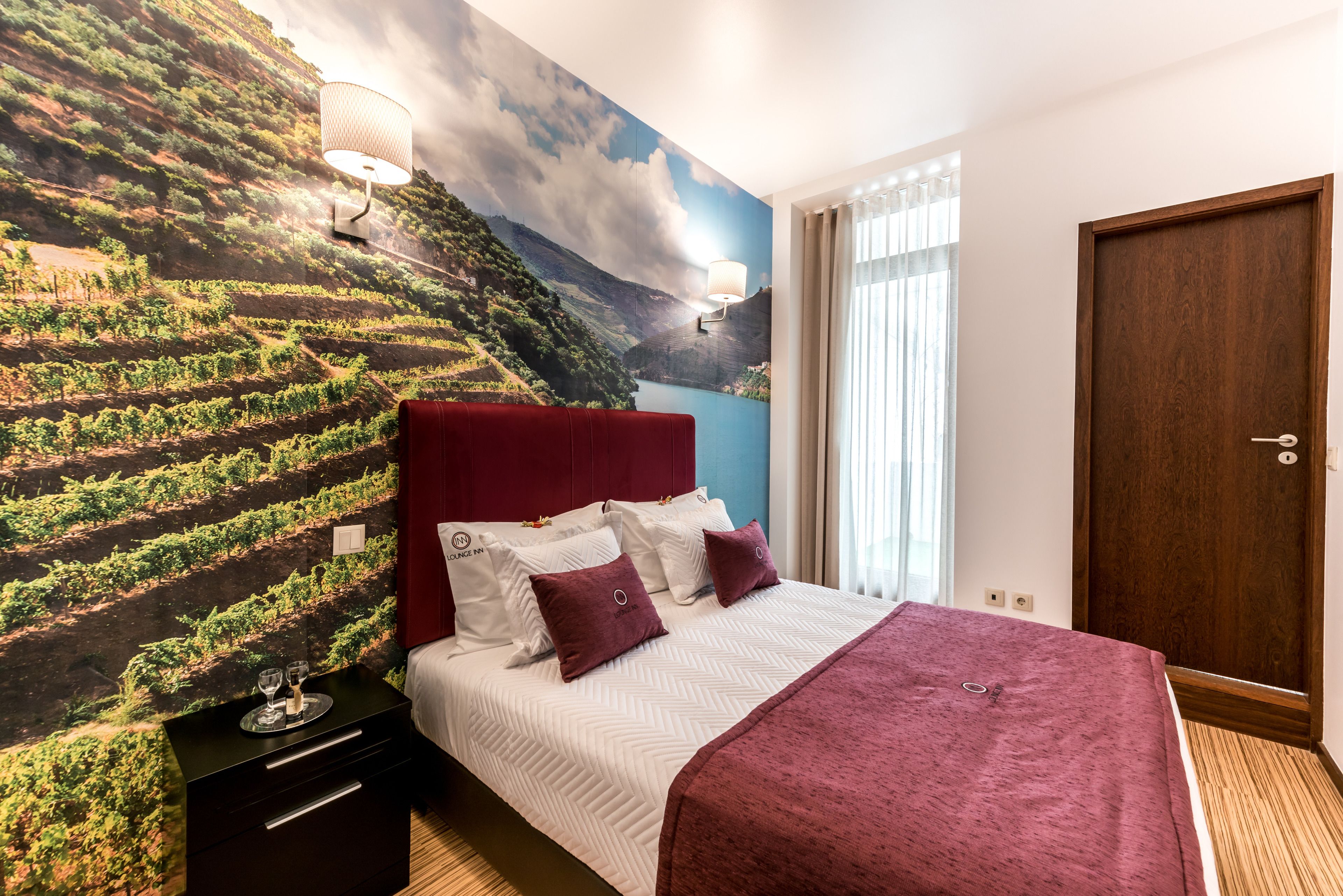 A hotel room with a bed, a nightstand, a red headboard, a wall mural of a vineyard, and a wooden door.