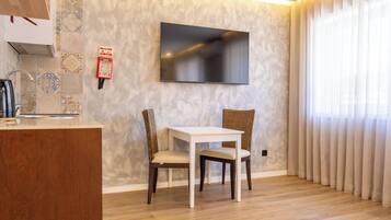 A modern living room with a flat-screen TV, a dining table, two chairs, a kitchen area with a kettle, and a patterned wall.
