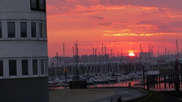 Un port de plaisance avec des bateaux amarrés, un coucher de soleil en arrière-plan et un bâtiment doté de nombreuses fenêtres.