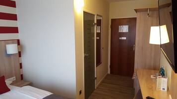 A hotel room with a bed, a desk with a lamp, a wardrobe, and a door with a sign.