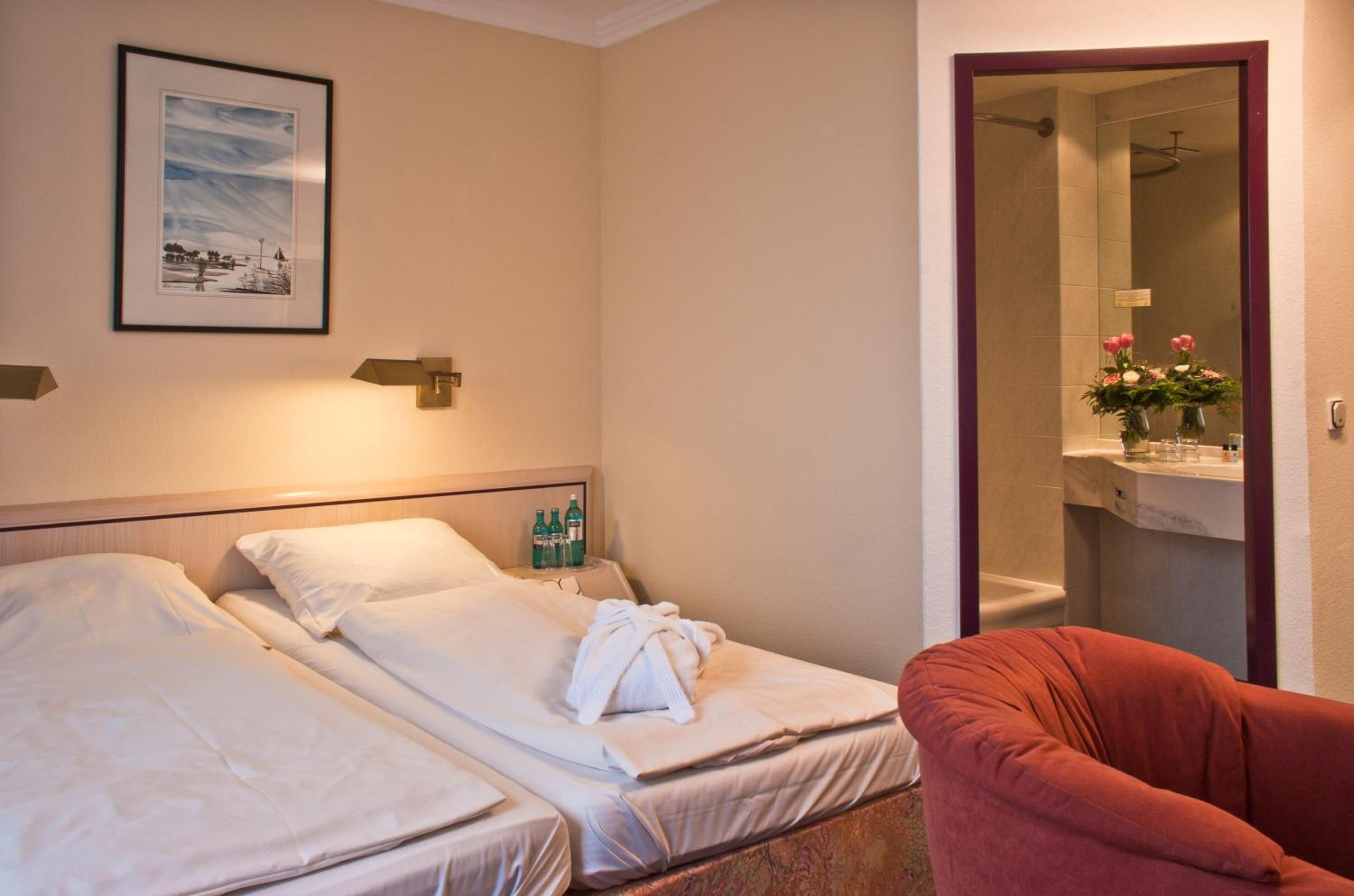 Habitación de hotel con dos camas, una silla roja, un cuadro en la pared y un baño visible a través de una puerta abierta.