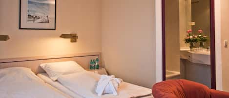 Habitación de hotel con dos camas, una silla roja, un cuadro en la pared y un baño visible a través de una puerta abierta.