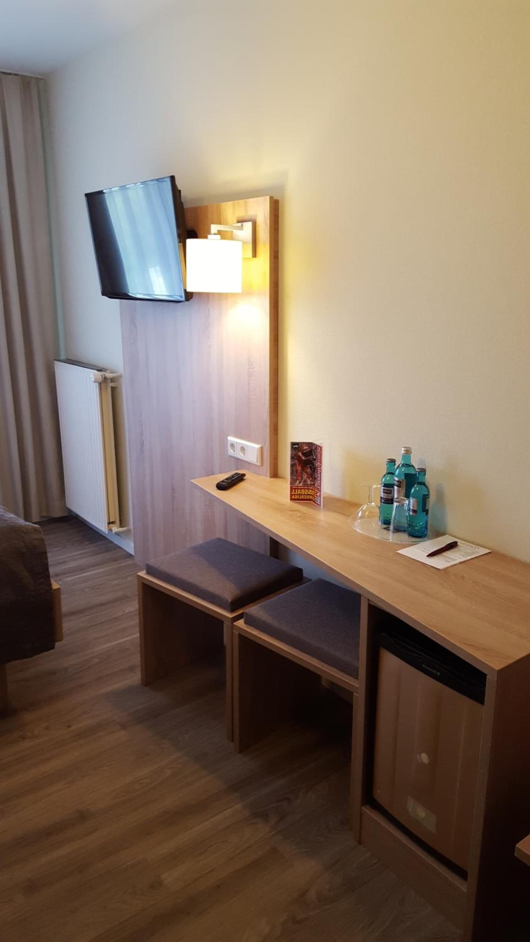Hotelkamer met een houten bureau, twee gestoffeerde bankjes, een flatscreen televisie aan de muur, een klein tafeltje met een afstandsbediening, een poster en een paar waterflesjes.