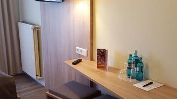 Hotelkamer met een houten bureau, twee gestoffeerde bankjes, een flatscreen televisie aan de muur, een klein tafeltje met een afstandsbediening, een poster en een paar waterflesjes.