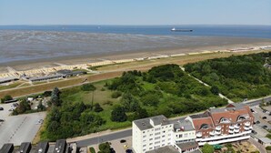 Een kustgebied met een strand, een groot schip en een gebouwencomplex met parkeerplaatsen en groen.