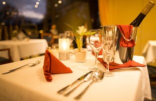 Une table à manger dressée avec une bouteille de champagne, deux verres à vin, une serviette rouge et des couverts.