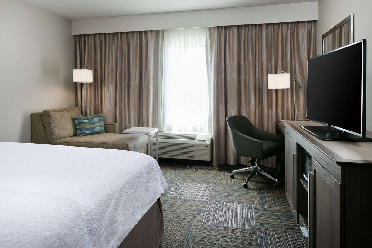 A hotel room with a bed, a desk, a chair, a TV, and a large window with curtains.