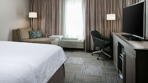 A hotel room with a bed, a desk, a chair, a TV, and a large window with curtains. - Hampton Inn & Suites Mason City (Mason City)