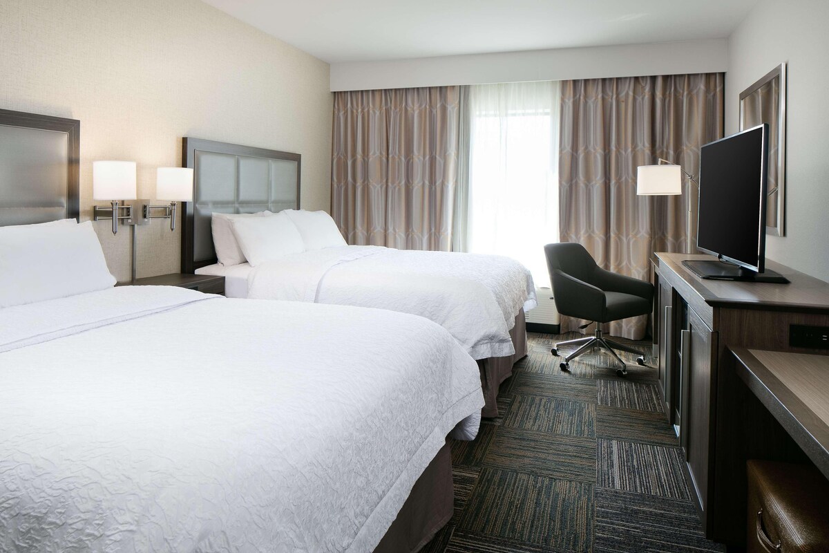 A hotel room with two beds, a desk with a television, a chair, and a large window with curtains.