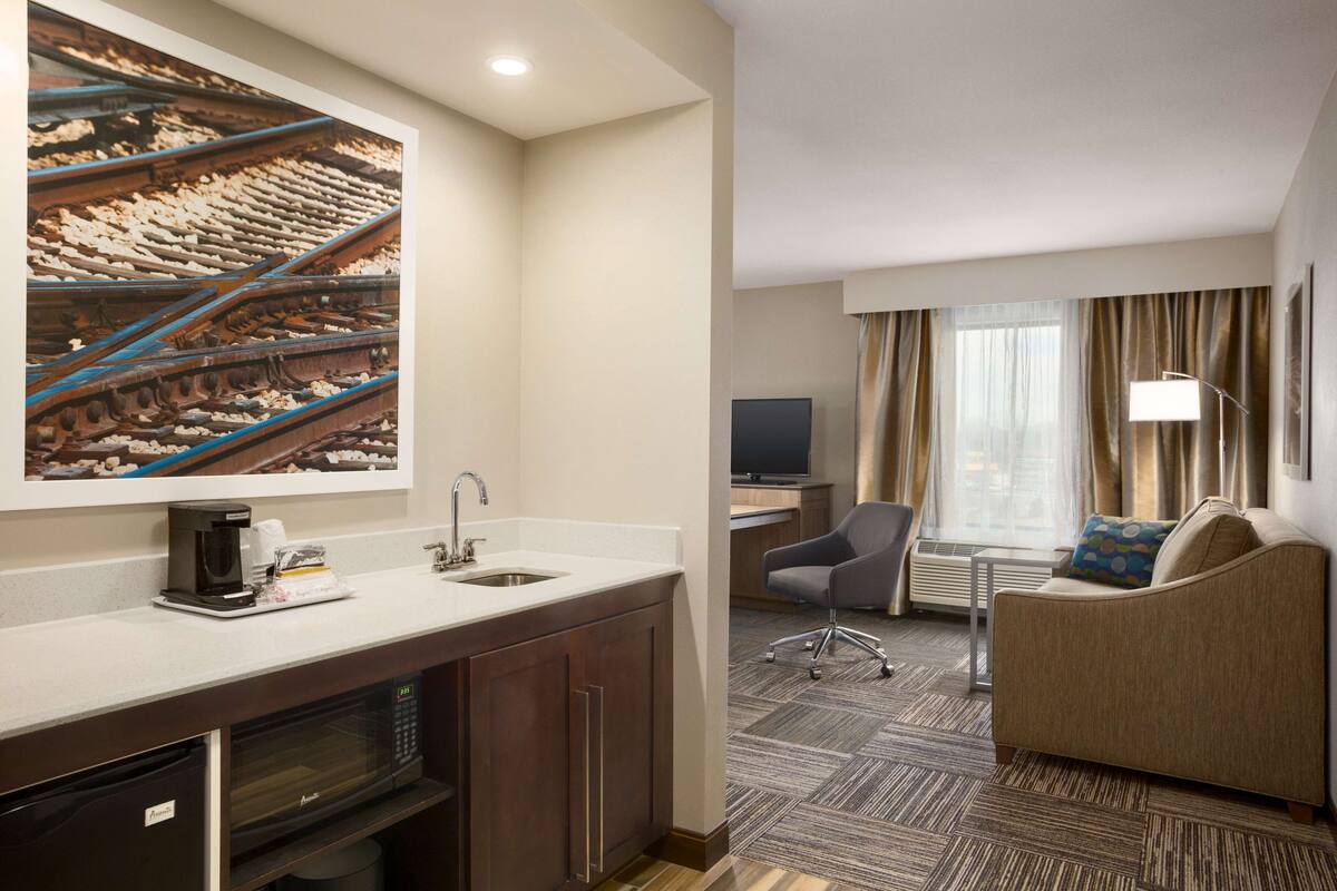 A hotel room with a kitchenette, a sofa, a TV, and a large window with curtains.