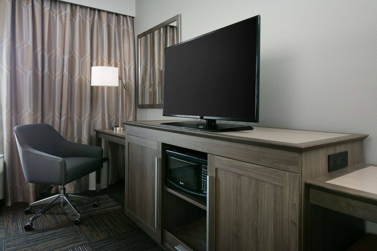 A hotel room with a flat-screen TV on a wooden desk, a modern chair, a microwave, and a lamp.
