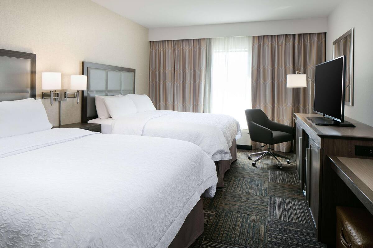 A hotel room with two beds, a desk with a television, a chair, and a large window with curtains.