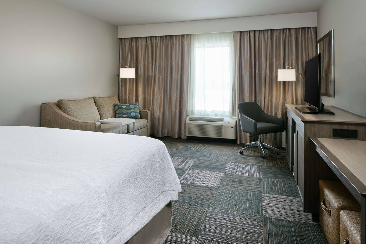 A hotel room with a large bed, a desk with a flat-screen TV, a chair, a desk lamp, and a window with curtains.