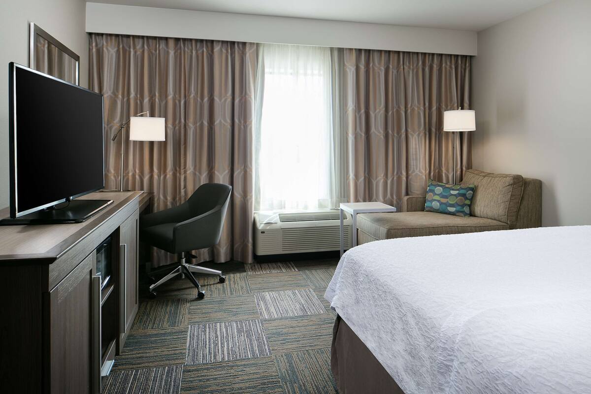 A hotel room with a bed, a desk with a TV, a chair, a sofa, and a window with curtains.