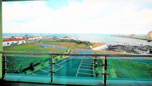 Une vue depuis un balcon donnant sur une zone côtière, avec un parking, de la végétation et des bâtiments visibles au loin.
