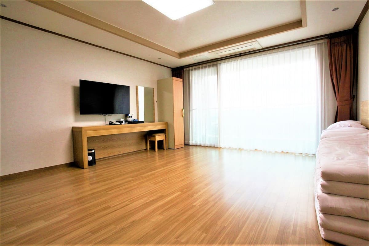 A hotel room with a wooden floor, a flat-screen TV mounted on the wall, a sofa, and a large window with curtains.