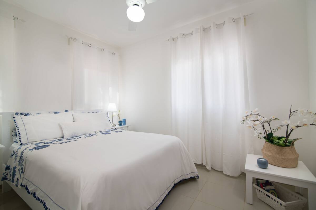 A white bedroom with a bed, curtains, a bedside table with a lamp, and a small plant on a nightstand.