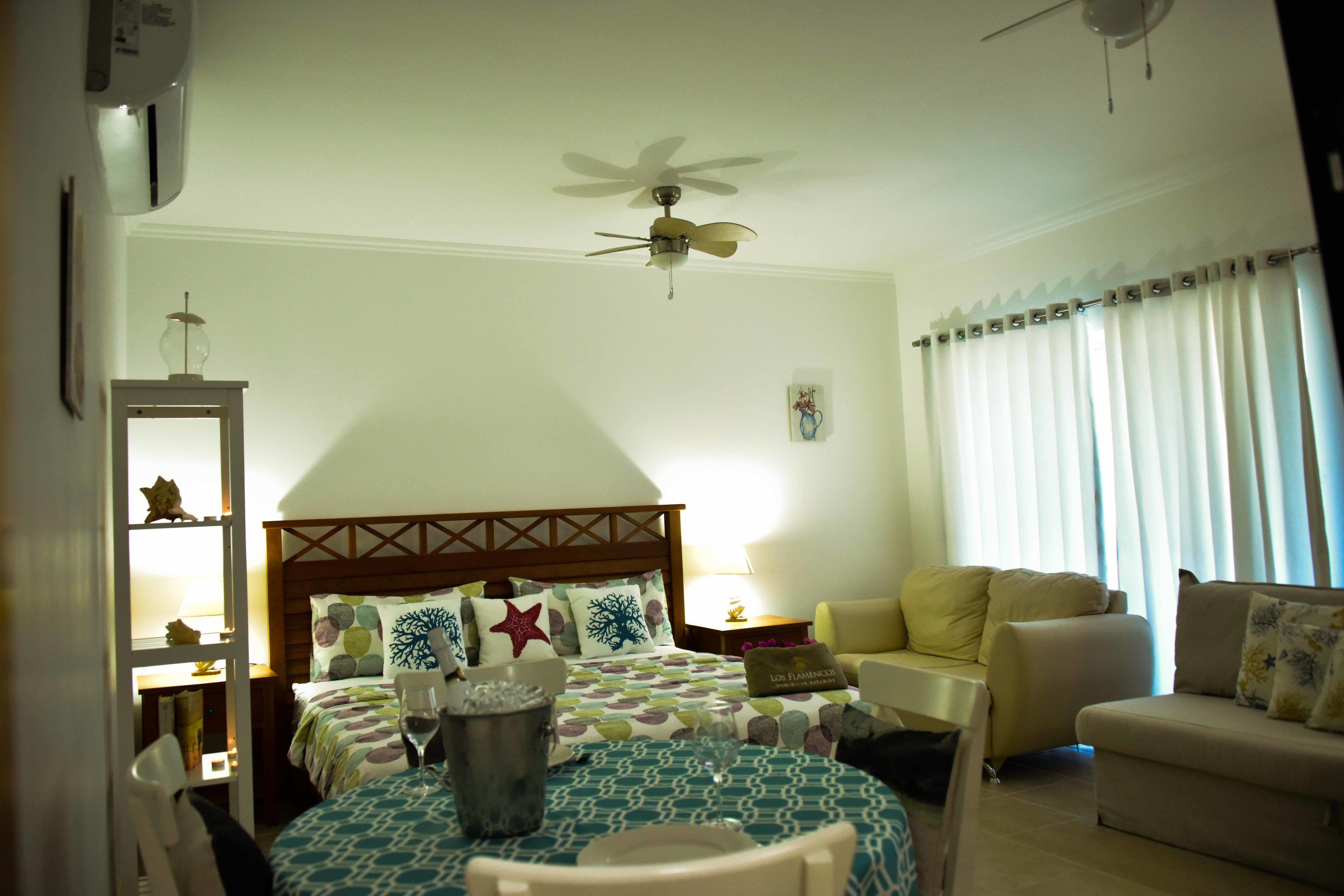 A hotel room with a bed, a dining table, a ceiling fan, a sofa, and a window with curtains.