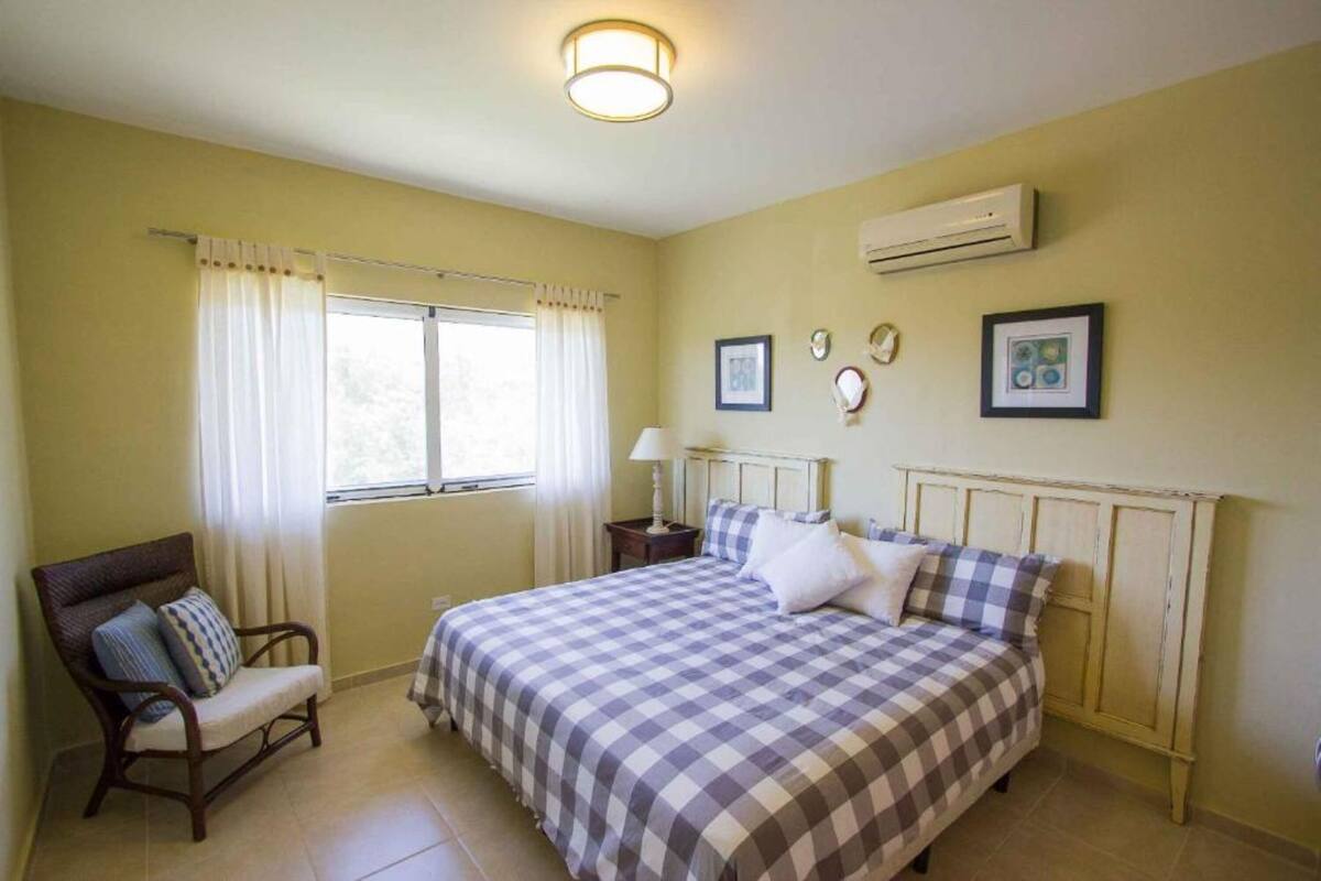 A bedroom with a bed, a chair, a window with curtains, an air conditioner, and wall decorations.