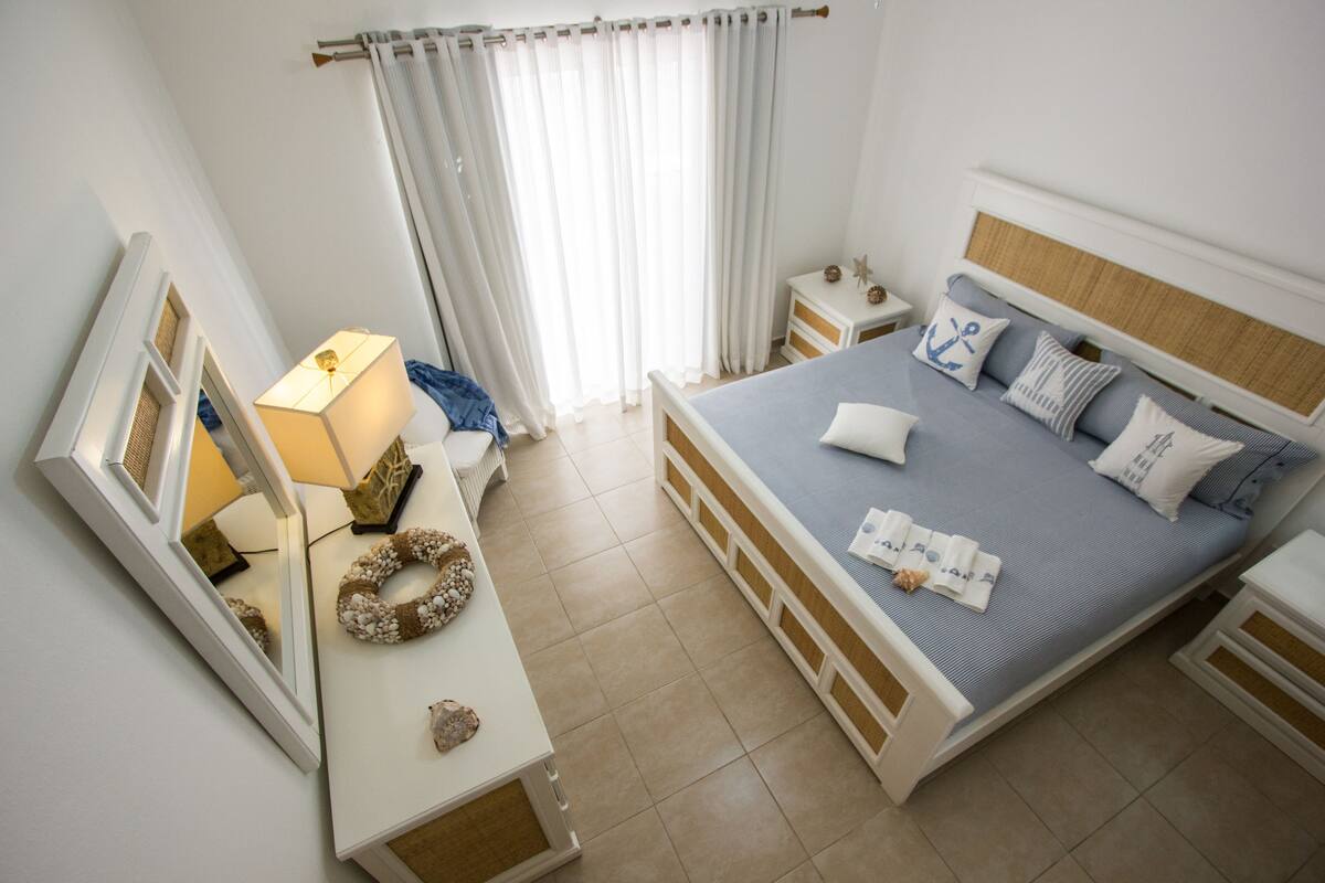 A neatly arranged bedroom with a bed, bedside tables, a dresser, a mirror, and a window with curtains.