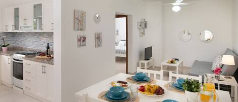 Una cocina moderna con armarios blancos, un comedor con vajilla azul y una sala de estar con televisor y sofá.