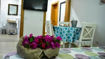 Une pièce avec une table recouverte d’une nappe à motifs, une chaise ornée d’une nappe bleue et blanche, et un petit panier tressé contenant des fleurs roses.